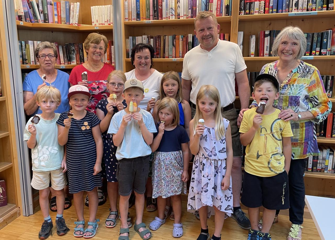 Kinder in der Bücherei stehend und ein Eis schleckend. Dahinter Erwachsene und Bücherregale