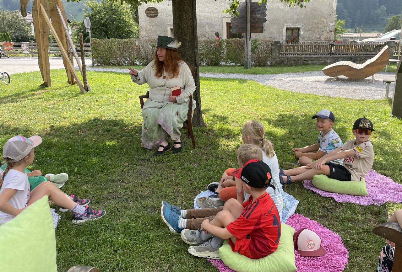 Kinder auf Picknickdecken auf der Wiese, Märchenerzählerin am Sessel vor ihnen sitzend.