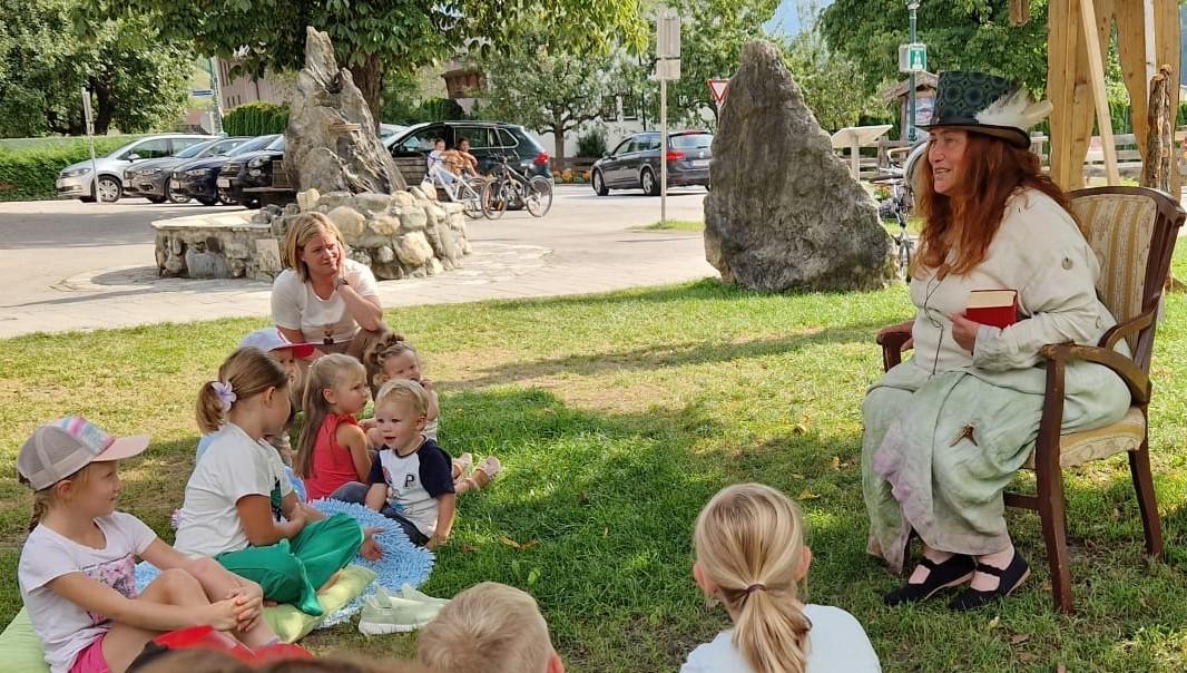 Märchenerzählerin am Sessel sitzend und Kinder davor auf Decken auf der Wiese sitzend