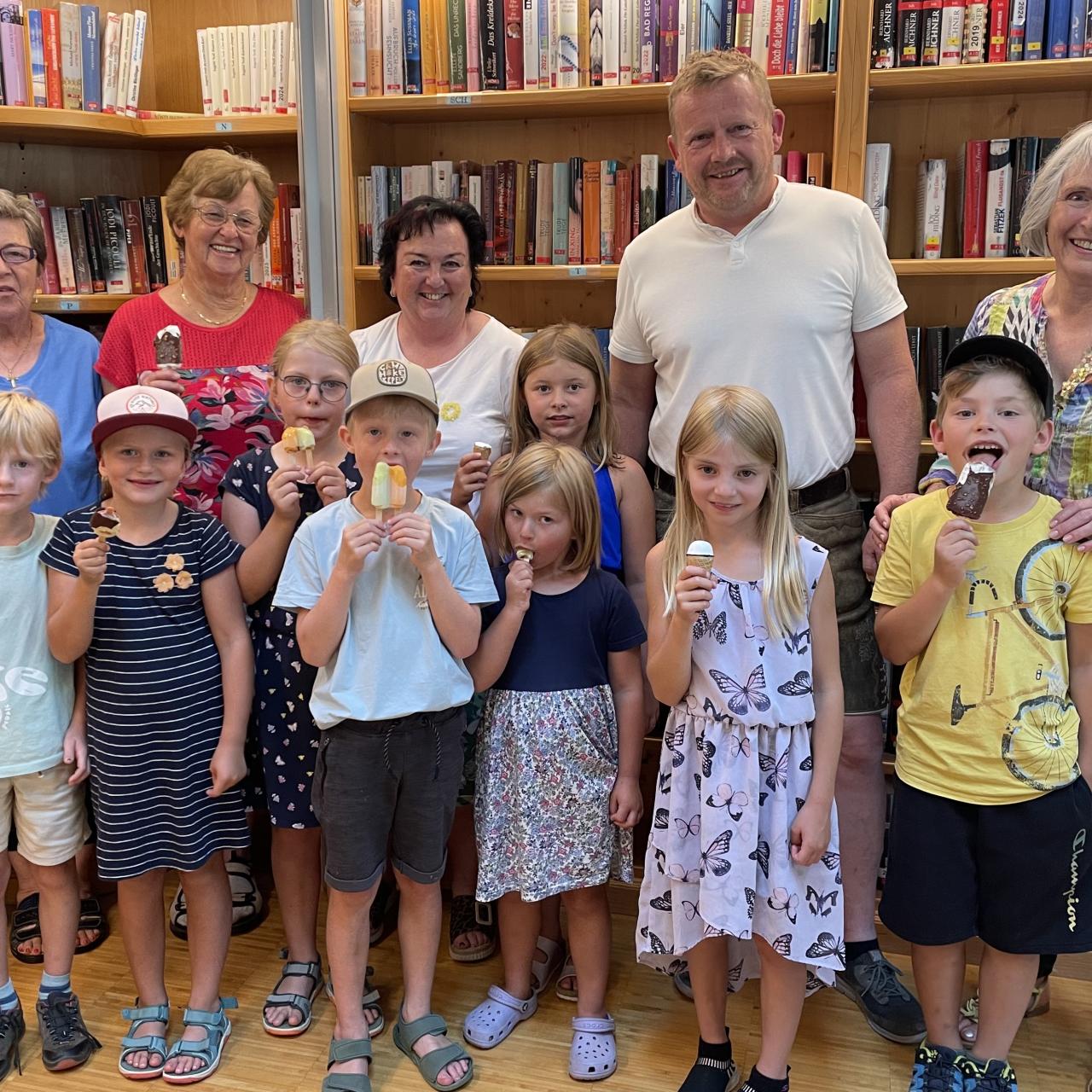 Kinder in der Bücherei stehend und ein Eis schleckend. Dahinter Erwachsene und Bücherregale